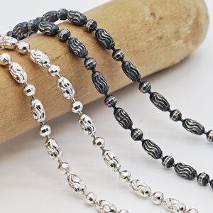 May include: Close-up of four beaded necklaces. Two are silver, and two are black. The silver necklaces have a mix of round and oval beads. The black necklaces have oval beads with a wavy design. The necklaces are on a wooden dowel.
