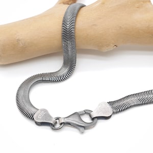 May include: A silver herringbone chain necklace with a lobster clasp. The necklace is displayed on a light brown wooden branch against a white background. The clasp is marked with "S925".