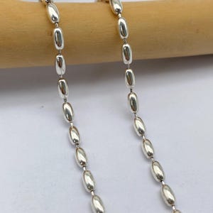 Può includere: Una collana a catena d'argento con maglie ovali. La catena è appoggiata su una superficie di legno.