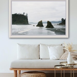 Peut inclure: Une photographie encadrée d'une plage rocheuse avec deux grandes formations rocheuses dans l'eau. Le ciel est couvert et l'eau est agitée. La photo est accrochée à un mur blanc au-dessus d'un canapé blanc avec un tapis tissé.