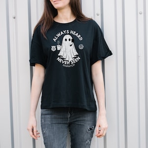 May include: A black t-shirt with a white graphic of a ghost wearing a headset and holding a cup of coffee. The text "Always Heard Never Seen Dispatch" is printed below the ghost.