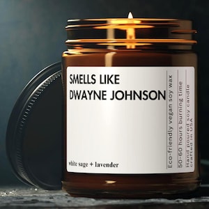 Può includere: Una candela accesa in un barattolo di vetro marrone con coperchio nero. L'etichetta recita "SMELLS LIKE DWAYNE JOHNSON" con "white sage + lavender" sotto. Il testo aggiuntivo include "Eco-friendly vegan soy wax".
