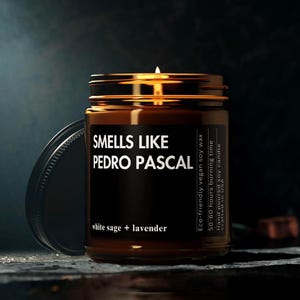 Può includere: Un barattolo di vetro marrone con un coperchio dorato contenente una candela. La candela è etichettata "Smells Like Pedro Pascal" e "white sage + lavender". L'etichetta indica anche che la candela è ecologica, in cera di soia vegana, ha un tempo di combustione di 50-60 ore, è realizzata a mano e prodotta negli Stati Uniti.