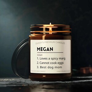 Puede incluir: Un tarro de vela de vidrio marrón con una tapa dorada. La etiqueta dice "DUCKY'S PREMIUM CANDLES" y "MEGAN" en negro. El texto debajo dice "nombre" y enumera tres descripciones humorísticas: "Le encanta un marg picante", "No puede cocinar huevos" y "La mejor mamá de perro". La parte inferior de la etiqueta dice "VEGAN SOY WAX CANDLE" y "HAND POURED IN USA".