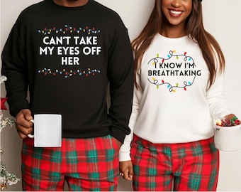 May include: Two matching sweatshirts, one black and one white, with festive text and fairy lights. The black sweatshirt reads "CAN'T TAKE MY EYES OFF HER", the white one says "I KNOW I'M BREATHTAKING". Both are worn with red and green plaid pyjama bottoms.