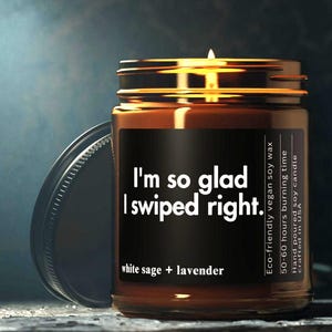 Könnte beinhalten: Eine brennende Sojawachskerze in einem Glas mit einem bronzefarbenen Deckel. Auf dem Etikett steht "I'm so glad I swiped right". Die Kerze duftet nach weißem Salbei und Lavendel. Der Text weist darauf hin, dass sie umweltfreundlich, vegan und in den USA handgegossen ist.
