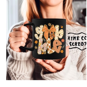 May include: Black ceramic mug with the word "SPOOKY HALE" in a fall-themed design. The design includes an orange bow, fall leaves, and a turkey. The mug is held by a person wearing a cream-colored sweater.