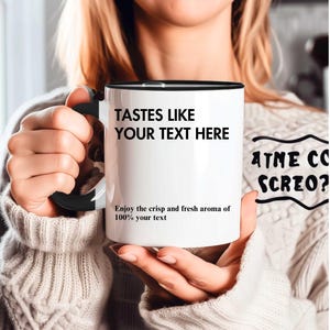 May include: White ceramic mug with a black interior and handle. The mug features the text "TASTES LIKE YOUR TEXT HERE" and "Enjoy the crisp and fresh aroma of 100% your text" in black font. The mug is held by a person.