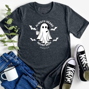May include: A dark gray t-shirt with a white ghost wearing a headset and holding a cup of coffee. The text on the shirt reads "Always Heard Never Seen Dispatch".