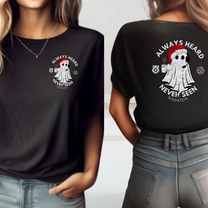 May include: Two women wearing black t-shirts with a white ghost graphic and the text "Always Heard Never Seen Dispatch". The ghost is wearing a Santa hat and holding a coffee cup.