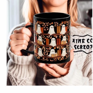 Peut inclure: Mug en céramique noire avec un motif de fantômes blancs sur des étagères et des feuilles d'automne. La tasse a une anse noire et est tenue par une personne portant un pull crème. Le mug porte le texte "ATNE CO SCREO?" sur le côté.