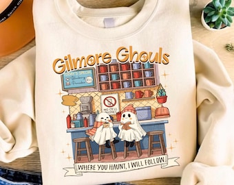 Könnte beinhalten: Cremefarbenes Sweatshirt mit dem Text "Gilmore Ghouls" und einer Diner-Szene-Grafik. Das Design zeigt Cartoon-Geister, ein "No Cell Phones"-Schild und den Satz "Where you haunt, I will follow" auf einem Banner.