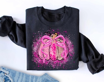 Op de afbeelding: Zwart sweatshirt met een roze pompoenontwerp, met een strik en roze lintaccenten. De pompoen heeft een luipaardprintpatroon en glitterdetails, met roze spettereffecten. Een thema voor borstkankerbewustzijn.