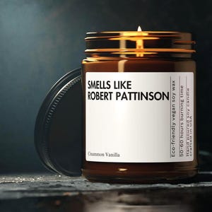 May include: A lit brown glass jar candle with a black lid. The label reads "Smells Like Robert Pattinson" and "Cinnamon Vanilla." The candle is eco-friendly vegan soy wax and hand-poured in the USA, with a 50-60 hour burn time.