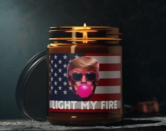 May include: A brown glass candle jar with a gold lid. The candle has a label with an American flag and a picture of a person wearing sunglasses and blowing a bubble. The label says "Light My Fire".