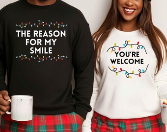 May include: A matching set of two black and white Christmas themed sweatshirts. The black sweatshirt has the text "The reason for my smile" with a string of colourful lights above and below the text. The white sweatshirt has the text "You're welcome" inside a circle of colourful lights.
