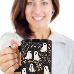 May include: Black coffee mug with a white ghost pattern on a black background. The ghosts are surrounded by small orange and white flowers.