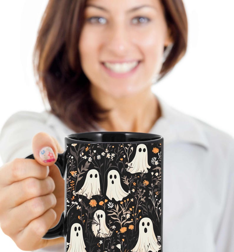 May include: Black coffee mug with a white ghost pattern on a black background. The ghosts are surrounded by small orange and white flowers.