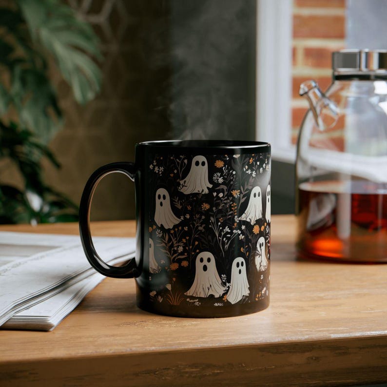 May include: Black ceramic mug with a white ghost pattern on a black background. The mug is filled with a hot beverage and steam is rising from the mug.