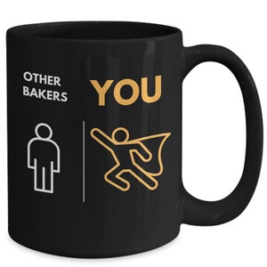May include: Black ceramic mug with the words "OTHER BAKERS" and a stick figure, contrasted with "YOU" and a superhero stick figure in gold. The mug has a large handle and a glossy finish.