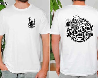 Può includere: T-shirt bianco con grafica nera di una mano di scheletro che fa un gesto rock and roll, con la parola "Fatherhood" sotto. Il retro presenta uno scheletro che beve birra con la scritta "Surviving Fatherhood One Beer At A Time".