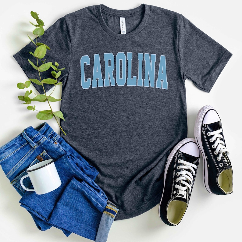 May include: A dark gray t-shirt with the word "CAROLINA" in light blue block letters. The shirt is paired with blue jeans, a white mug, and black canvas sneakers with white laces. A sprig of greenery adds a natural touch to the arrangement.