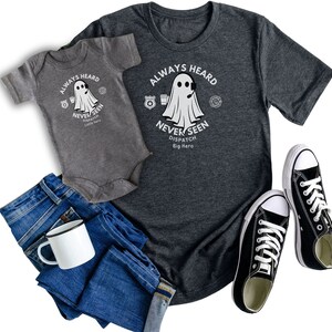 以下が含まれることがあります： グレーのTシャツとグレーのロンパースに、幽霊のグラフィックと「Always Heard Never Seen Dispatch」というテキストがプリントされています。その下に「Little Hero」と「Big Hero」という文字があります。