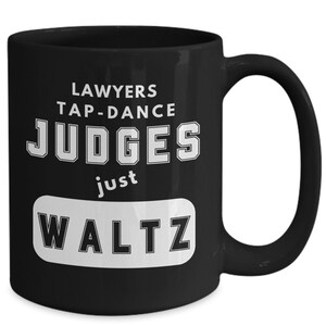 May include: Black coffee mug with white text that reads "Lawyers tap-dance Judges just waltz".