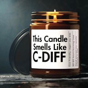 May include: A brown glass jar candle with a lit wick. The label reads "This Candle Smells Like C-DIFF." Additional text includes "Eco-friendly vegan soy wax" and "50-60 hours burning time."