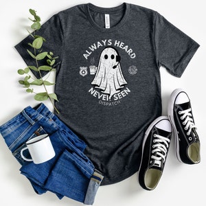May include: A dark grey t-shirt with a white distressed graphic of a ghost wearing a headset and holding a coffee cup. The text "Always Heard Never Seen Dispatch" is printed below the ghost.