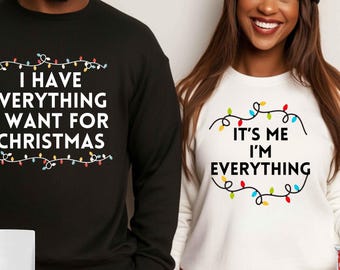 May include: Two Christmas-themed sweatshirts. One black sweatshirt says "I have everything I want for Christmas" with a string of colourful lights. The other white sweatshirt says "It's me I'm everything" with a string of colourful lights.