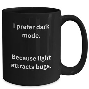 以下が含まれることがあります： 黒いセラミックマグカップ、湾曲したハンドル付き。 マグカップには、白い文字で「私はダークモードが好きです。光は虫を引き寄せるからです。」と書かれています。