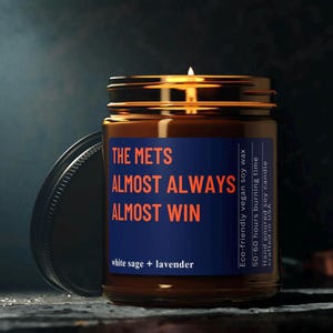 May include: A brown glass candle jar with a blue label that reads "THE METS ALMOST ALWAYS ALMOST WIN" and "white sage + lavender". The label also includes information about the candle, such as the type of wax, burn time, and where it was made.