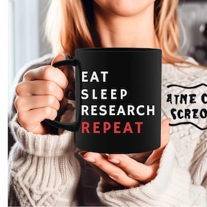 May include: Black coffee mug with white text that reads "Eat Sleep Research Repeat" in a vertical list. The text "Repeat" is in red. The mug is being held by a person wearing a black sweater.