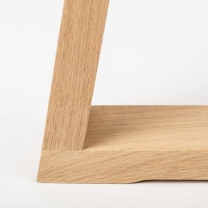 May include: A light-colored wooden shelf featuring a visible natural grain pattern. The structure consists of a horizontal flat board joined to a vertical support piece at an angled connection, showcasing the light tan wood texture and finish.