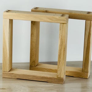 May include: Two square-shaped furniture legs constructed from light-colored natural wood with a visible grain pattern. Each leg features a sturdy frame design with four pre-drilled circular holes at the top corners for mounting purposes.