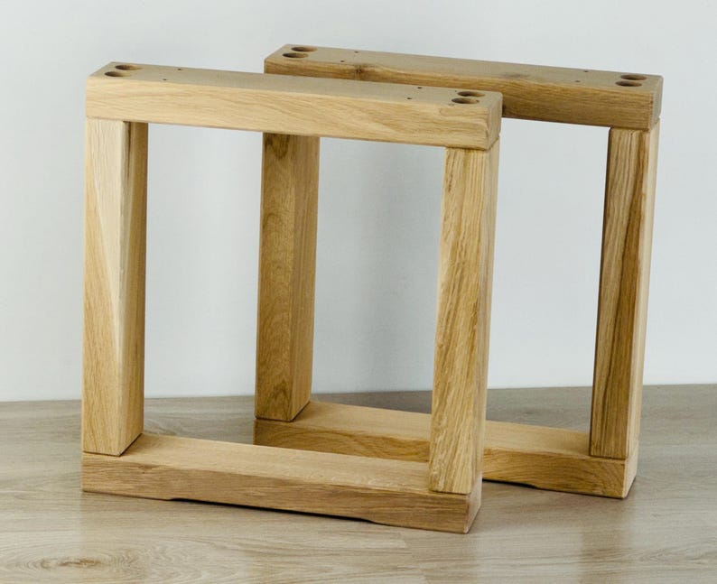 May include: Two square-shaped furniture legs constructed from light-colored natural wood with a visible grain pattern. Each leg features a sturdy frame design with four pre-drilled circular holes at the top corners for mounting purposes.