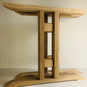 Puede incluir: Una base de mesa de madera clara con una superficie superior rectangular sostenida por dos pilares verticales conectados por dos travesa&ntilde;os horizontales, que descansa sobre una base ancha y plana con bordes angulados y veta natural.