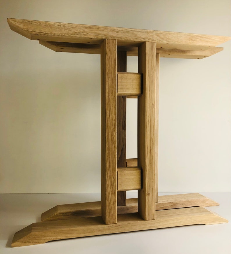 Puede incluir: Una base de mesa de madera clara con una superficie superior rectangular sostenida por dos pilares verticales conectados por dos travesa&ntilde;os horizontales, que descansa sobre una base ancha y plana con bordes angulados y veta natural.