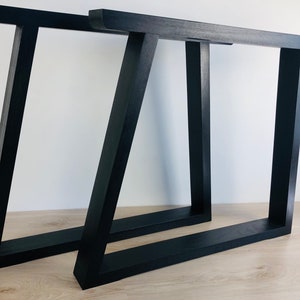 May include: Two black table legs with a trapezoidal frame design. Each leg features a horizontal top bar, a horizontal base bar, and two vertical supports, with one support angled to create a distinct geometric shape. The finish is matte black.