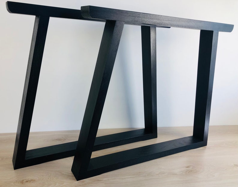 May include: Two black table legs with a trapezoidal frame design. Each leg features a horizontal top bar, a horizontal base bar, and two vertical supports, with one support angled to create a distinct geometric shape. The finish is matte black.
