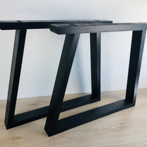 May include: Two black wooden table legs with a trapezoidal frame design. Each leg features a horizontal base, a vertical side, and an angled side, with pre-drilled mounting holes visible on the top horizontal support beam of each structure.