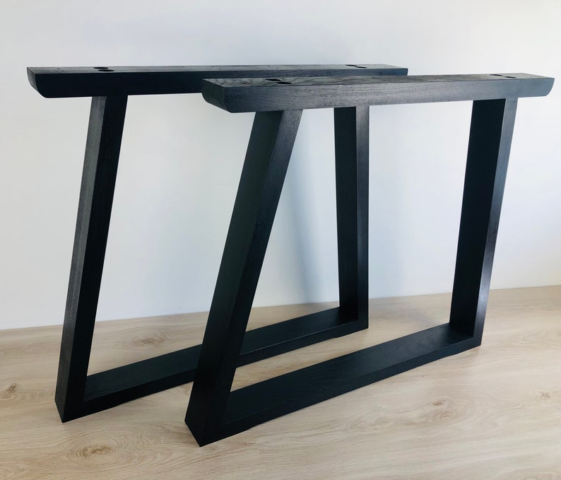 May include: Two black wooden table legs with a trapezoidal frame design. Each leg features a horizontal base, a vertical side, and an angled side, with pre-drilled mounting holes visible on the top horizontal support beam of each structure.