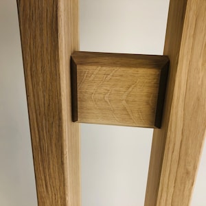 Puede incluir: Una estructura de madera de color marr&oacute;n claro con dos postes verticales conectados por un bloque rectangular horizontal. La madera muestra un patr&oacute;n de veta natural con radios medulares visibles y un acabado liso en la pieza central.