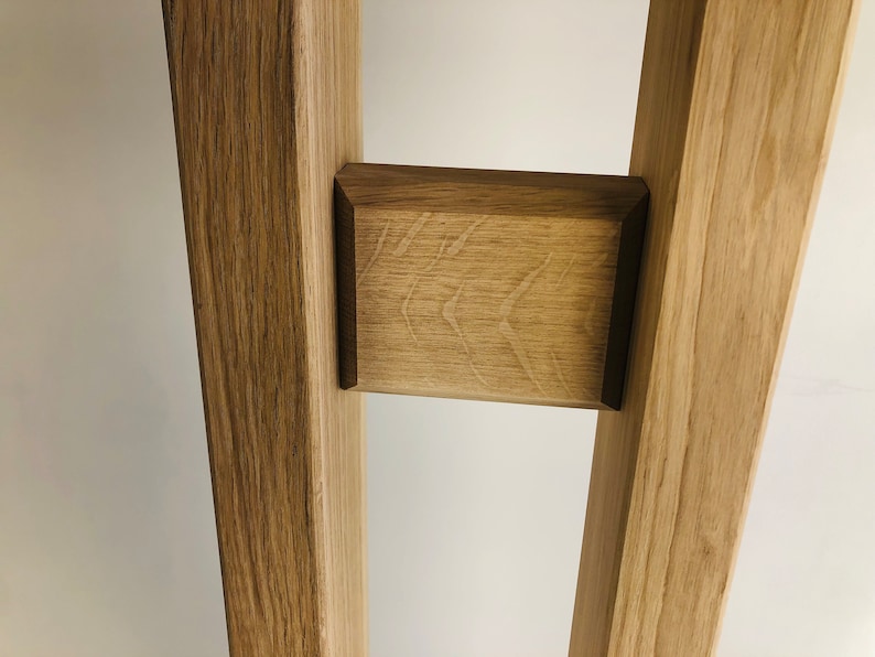 Puede incluir: Una estructura de madera de color marr&oacute;n claro con dos postes verticales conectados por un bloque rectangular horizontal. La madera muestra un patr&oacute;n de veta natural con radios medulares visibles y un acabado liso en la pieza central.