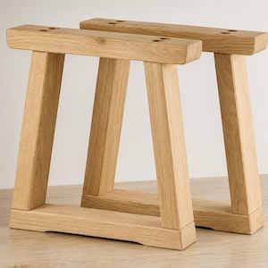 Può includere: Una coppia di gambe per tavolo in legno chiaro con un design a telaio trapezoidale. Ogni gamba è composta da due supporti verticali angolati collegati da una base orizzontale e da una piastra di montaggio superiore con fori circolari preforati.