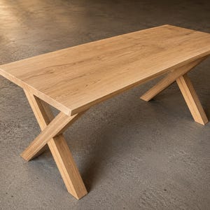 Könnte beinhalten: Ein rechteckiger Esstisch aus hellem Holz mit sichtbarer Maserung. Der Tisch verfügt über eine flache, glatte Oberfläche, die von zwei sich kreuzenden Beinpaaren getragen wird, welche an beiden Enden des Möbelstücks eine X-Form bilden.