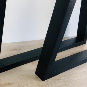 May include: Two black table legs constructed from solid wood with a visible grain texture. Each leg features a rectangular base frame connected to an angled vertical support, creating a geometric, open-frame structure with clean, sharp edges.