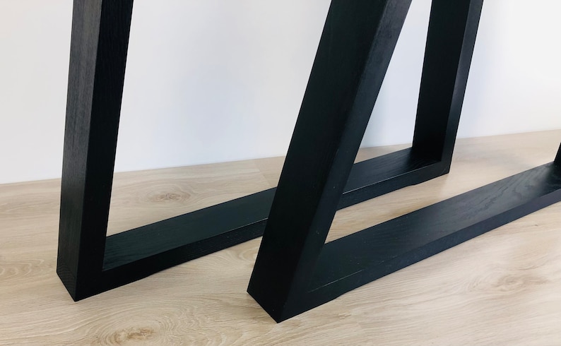 May include: Two black table legs constructed from solid wood with a visible grain texture. Each leg features a rectangular base frame connected to an angled vertical support, creating a geometric, open-frame structure with clean, sharp edges.