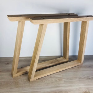 May include: Two light-colored wooden table legs featuring a trapezoidal frame design. Each leg has a flat top mounting plate with pre-drilled holes and a slanted vertical support connected to a horizontal base piece, showcasing a natural wood grain.